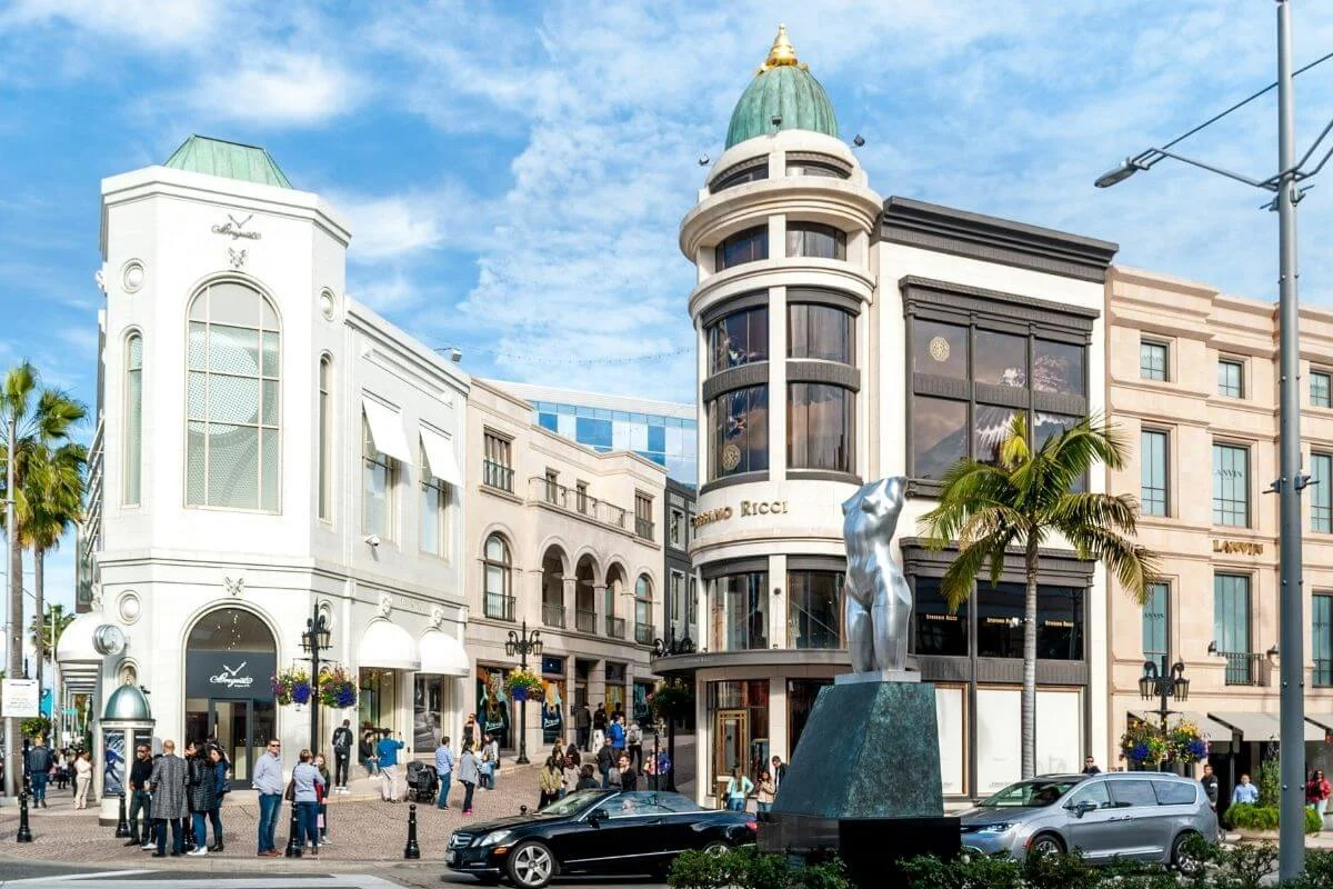 Luxury storefronts and shoppers along Rodeo Drive in Beverly Hills featuring designer boutiques, palm trees, and the iconic public sculpture at the intersection on a sunny day