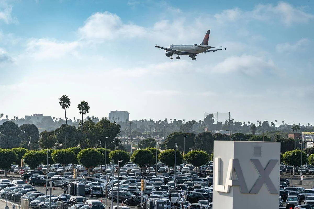 Commercial airplane approaching landing above Los Angeles International Airport with the LAX sign and parking lot visible below.
