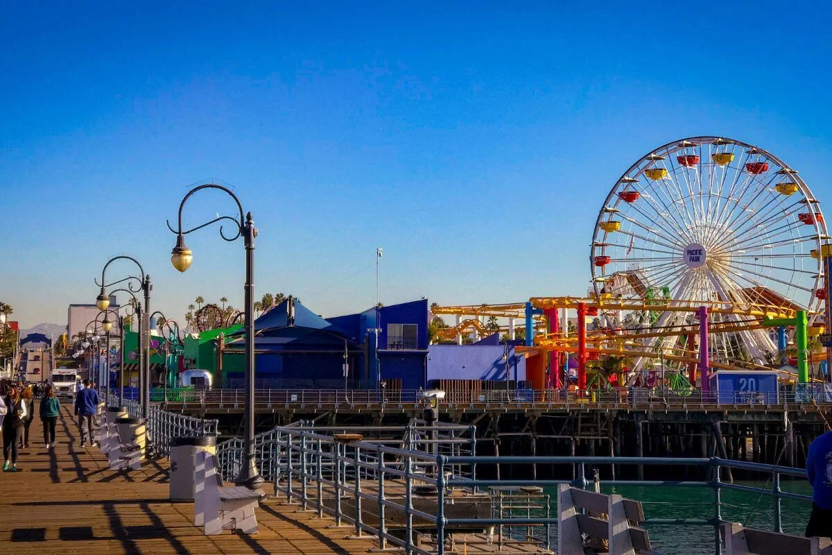 A sunny daytime view of Santa Monica Pier featuring a colorful Ferris wheel, amusement rides, and people walking along the wooden boardwalk by the ocean.