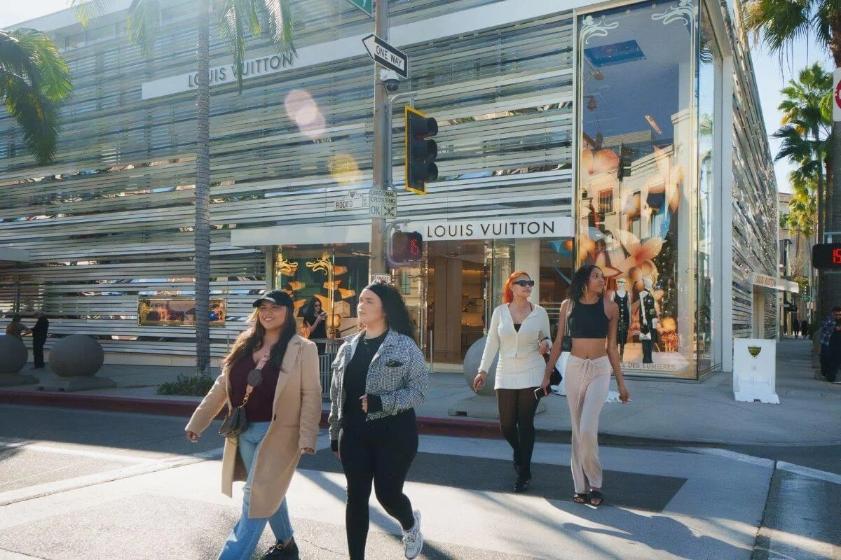 People walking past a Louis Vuitton store on Rodeo Drive in Beverly Hills, surrounded by palm trees and luxury storefronts on a sunny day.