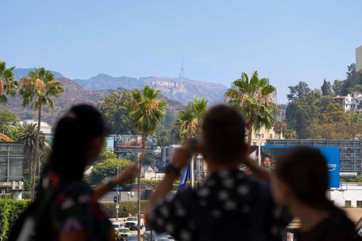 Visitors taking photos of the Hollywood Sign in the hills of Los Angeles with palm trees and city streets in the foreground.