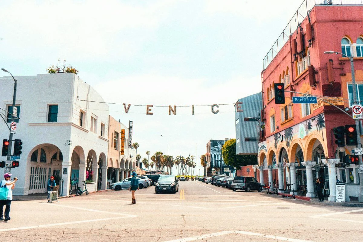 A sunny street view of Venice Beach with the iconic “VENICE” sign hanging above the road, colorful buildings on both sides, and palm trees visible in the distance.