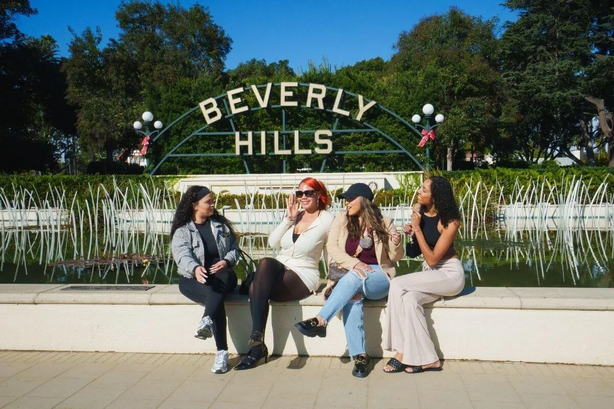 A group of friends sitting and posing in front of the iconic Beverly Hills sign with a fountain and greenery in the background on a sunny day.