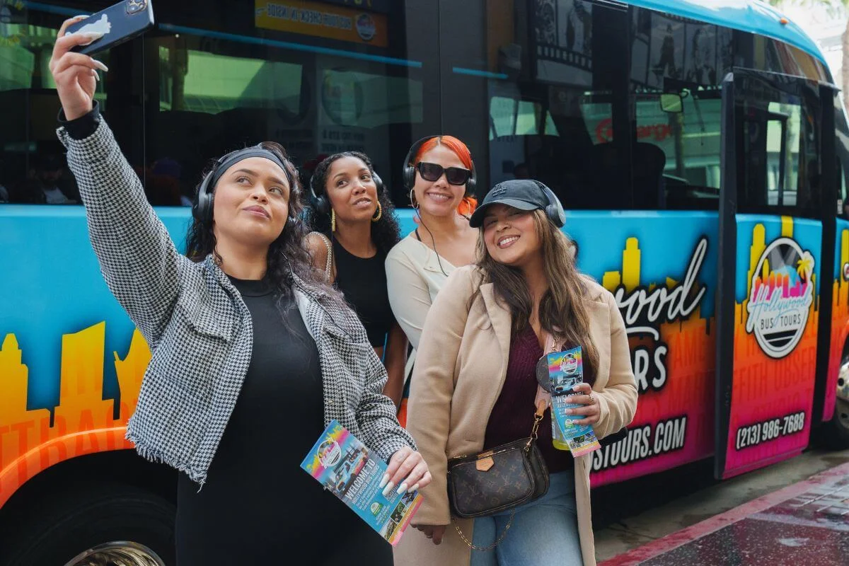 Group of friends wearing headphones taking a selfie beside a Hollywood Bus Tours sightseeing bus in Los Angeles
