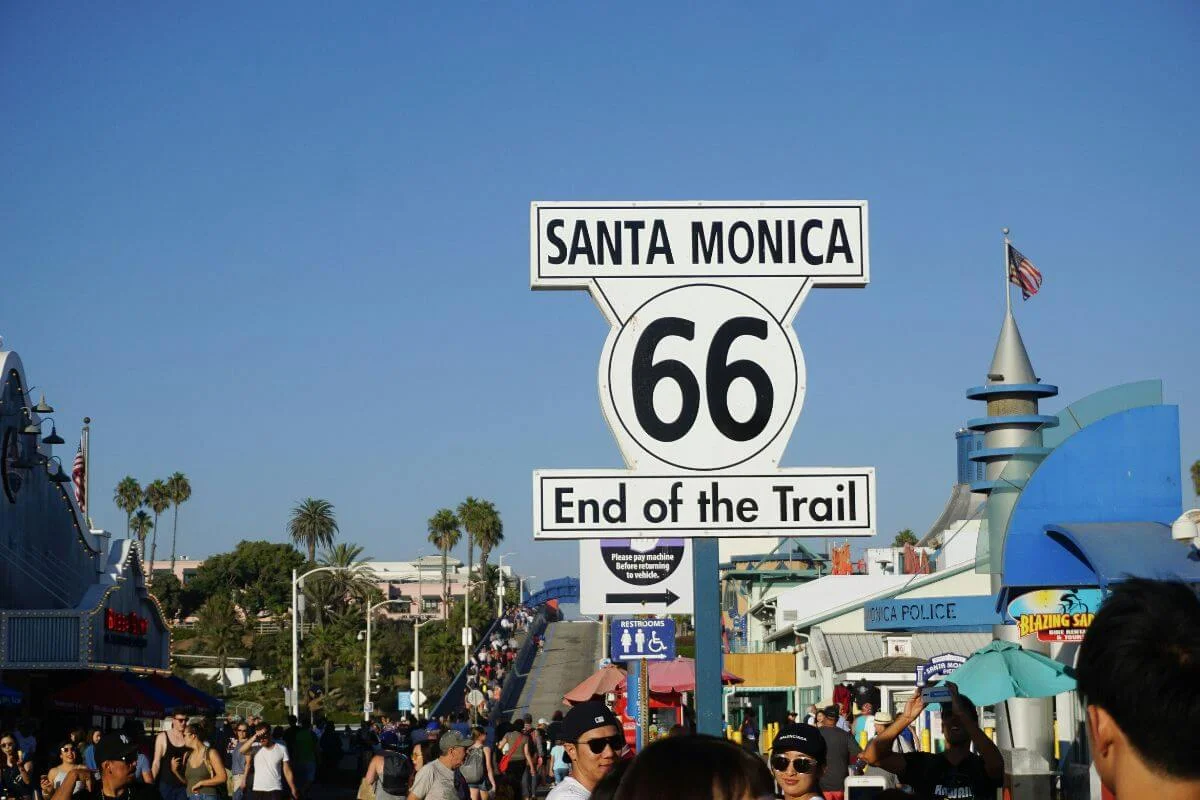 A crowd of people gathered around the iconic Route 66 “End of the Trail” sign at Santa Monica, with shops, palm trees, and a busy pier atmosphere in the background.