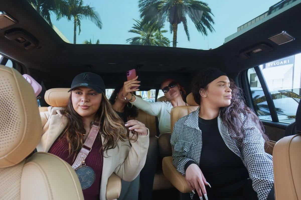 A group of friends riding inside a vehicle, taking photos and enjoying views of Beverly Hills with palm trees and luxury stores visible outside.