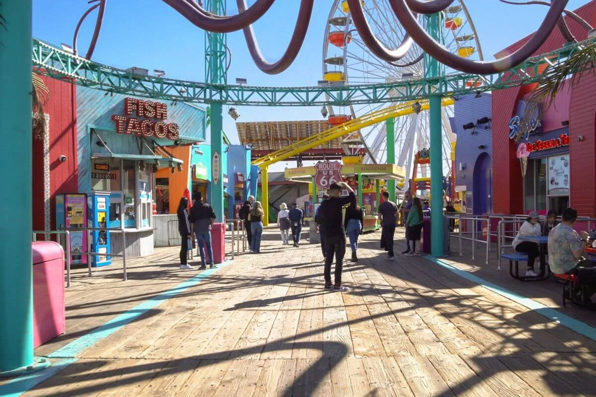 People walking along the colorful Santa Monica Pier boardwalk with food stalls, shops, and amusement rides including a Ferris wheel in the background.