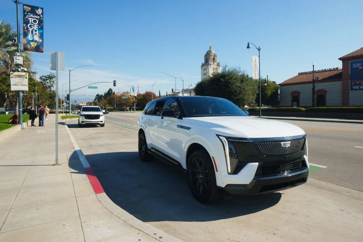 A white luxury SUV driving along a sunny Los Angeles street with palm trees, buildings, and light traffic in the background.