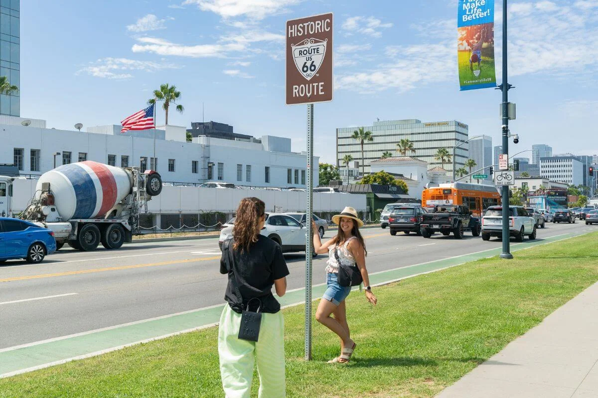 Visitor posing for a photo beside a Historic Route 66 sign along Santa Monica Boulevard in Los Angeles.