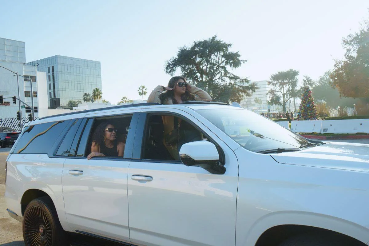 Two women enjoying a Los Angeles sightseeing ride in a white SUV with palm trees and city buildings in the background.