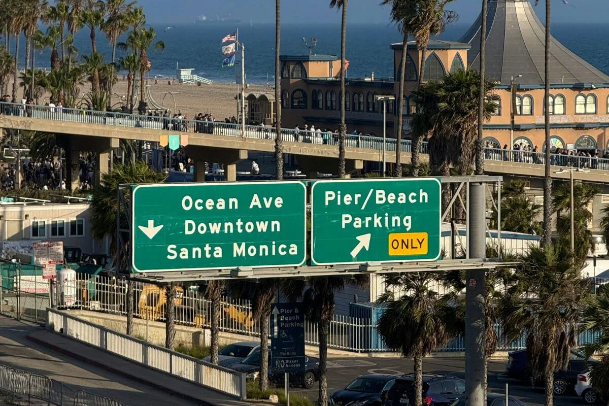 Green road signs pointing to Ocean Avenue, downtown Santa Monica, and pier beach parking, with palm trees, buildings, and the ocean visible in the background.