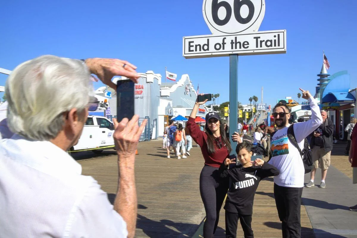 A group of tourists posing for a photo at the Route 66 “End of the Trail” sign on Santa Monica Pier while another person takes their picture.