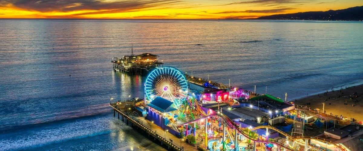 A vibrant aerial view of Santa Monica Pier at sunset, featuring a brightly lit Ferris wheel and amusement rides extending over the ocean with colorful lights reflecting on the water.