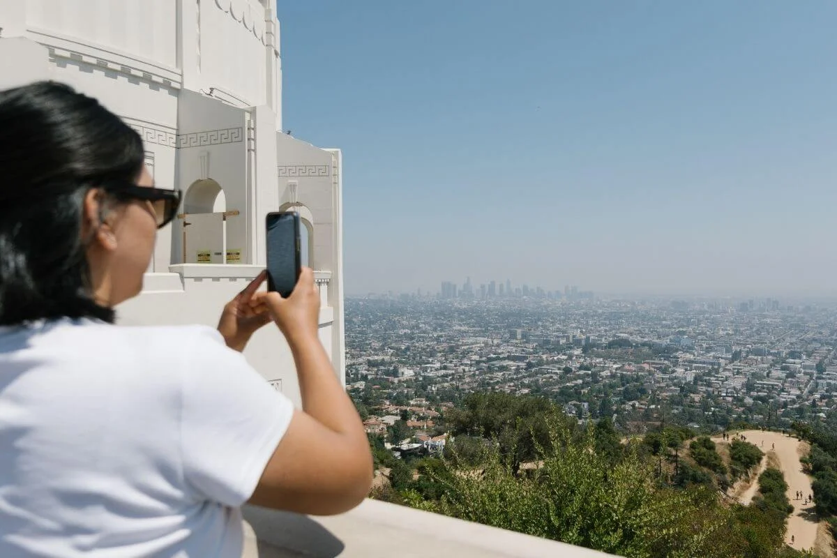 Person taking a photo of the Los Angeles skyline from a scenic viewpoint near Griffith Observatory, overlooking the city and surrounding hills.