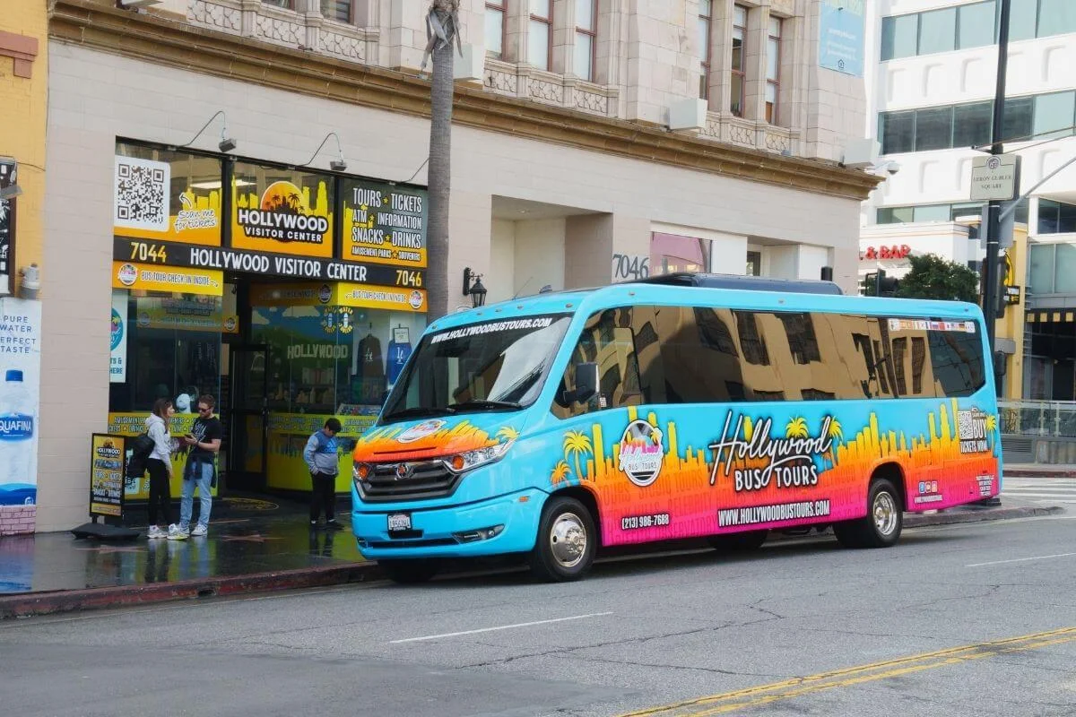 Colorful Hollywood Bus Tours vehicle parked in front of the Hollywood Visitor Center as people stand nearby on the sidewalk in Los Angeles.