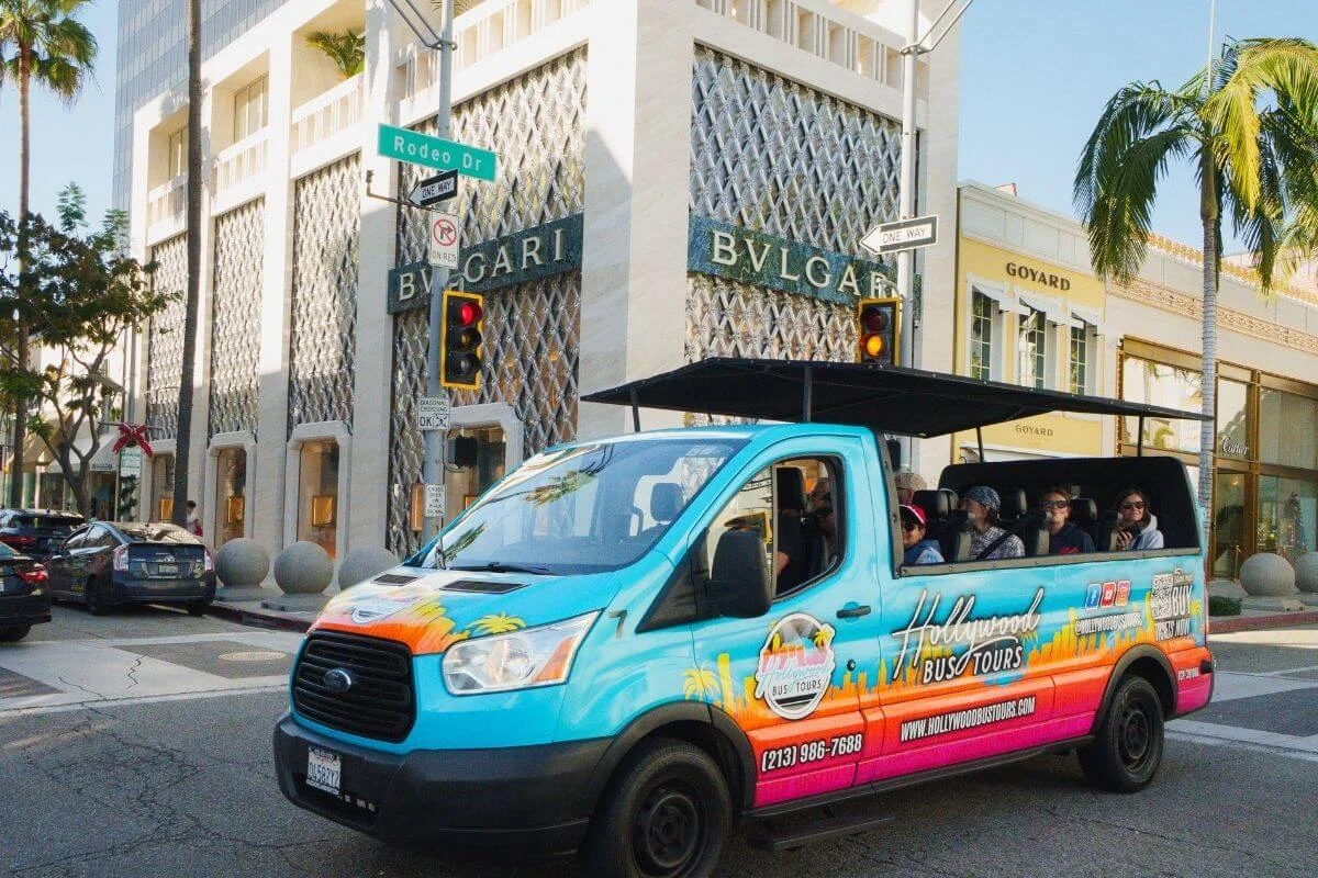 Colorful Hollywood Bus Tours van with passengers driving past luxury stores on Rodeo Drive in Beverly Hills, Los Angeles.