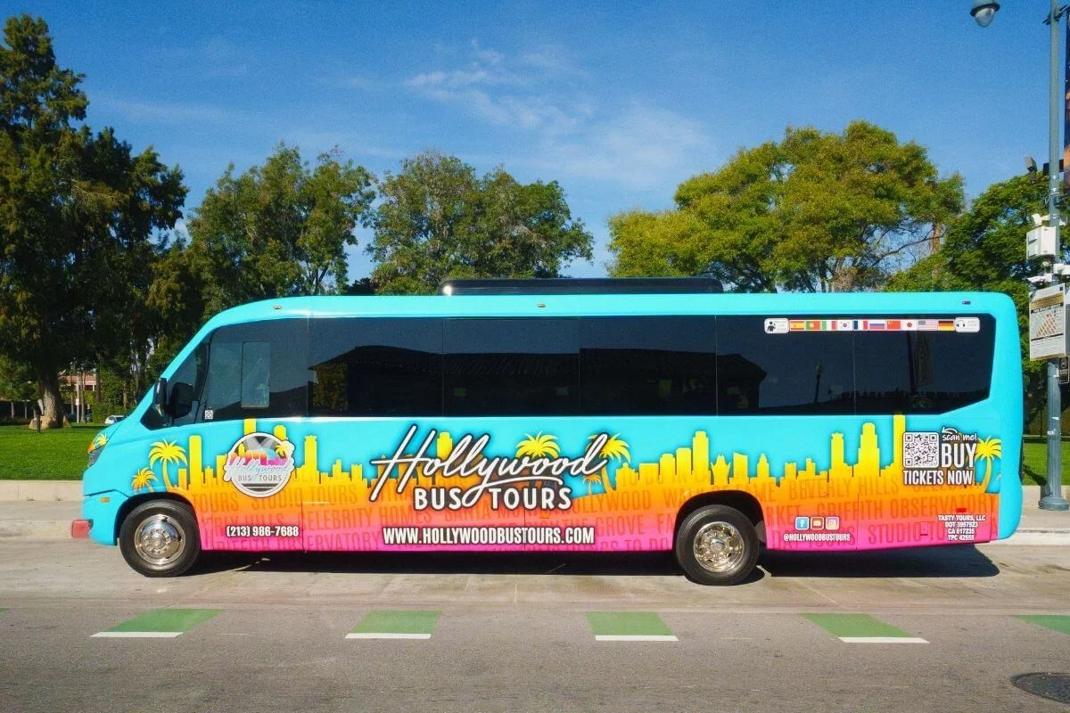 Brightly colored Hollywood Bus Tours shuttle parked along a sunny Los Angeles street, featuring skyline graphics and branding on the vehicle exterior.