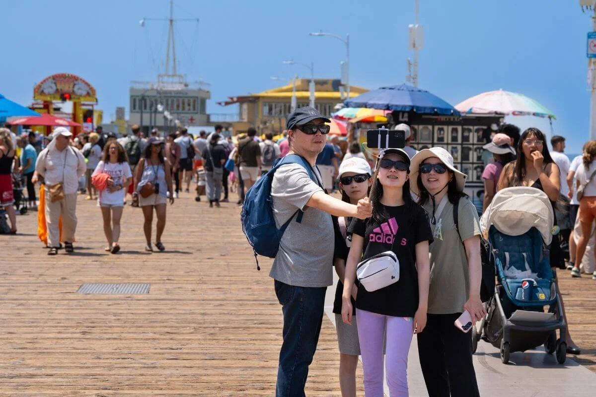 Family using a selfie stick while walking along the busy Santa Monica Pier, surrounded by tourists, shops, and oceanfront attractions in Los Angeles.