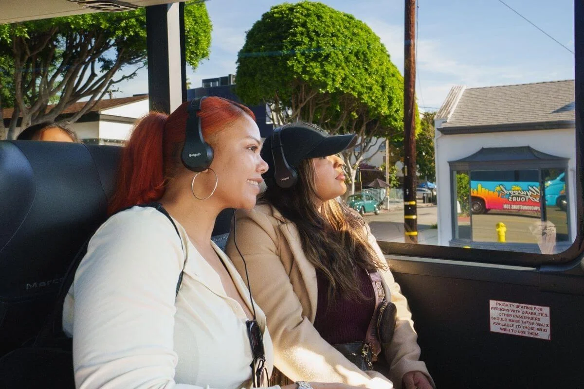 Two women wearing headsets smiling and looking out the window while riding a Hollywood bus tour through a sunny Los Angeles neighborhood.