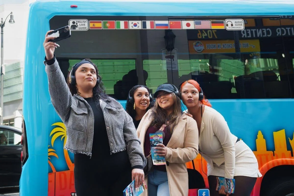 Group of women taking a selfie in front of a colorful Hollywood bus tour vehicle, holding brochures and enjoying a sightseeing experience in Los Angeles.