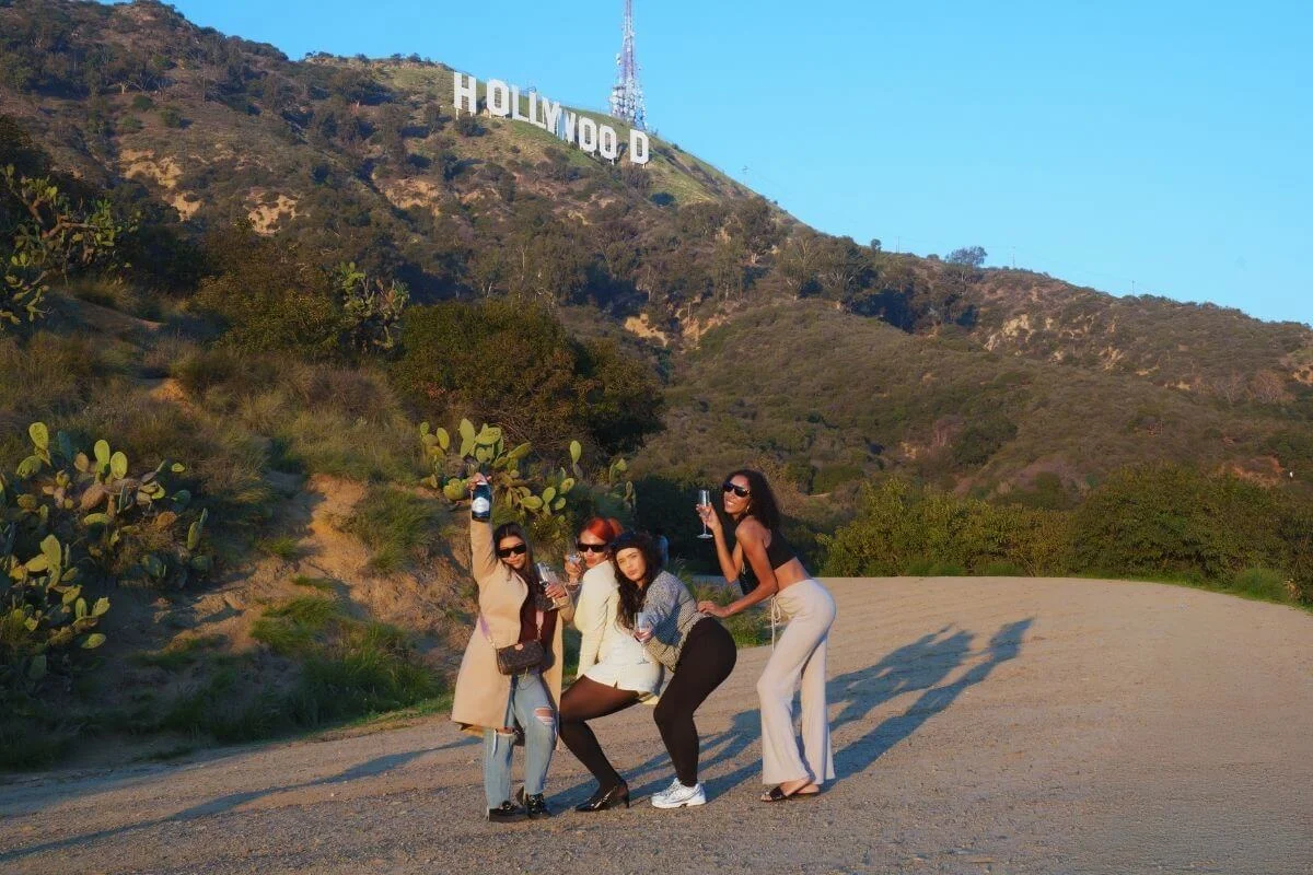 Group of friends posing and having fun on a scenic trail with the Hollywood sign visible on the hillside in Los Angeles during golden hour.