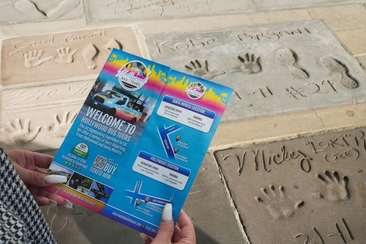 Visitor holding a Hollywood Bus Tours brochure over celebrity handprints at the TCL Chinese Theatre on the Hollywood Walk of Fame in Los Angeles.