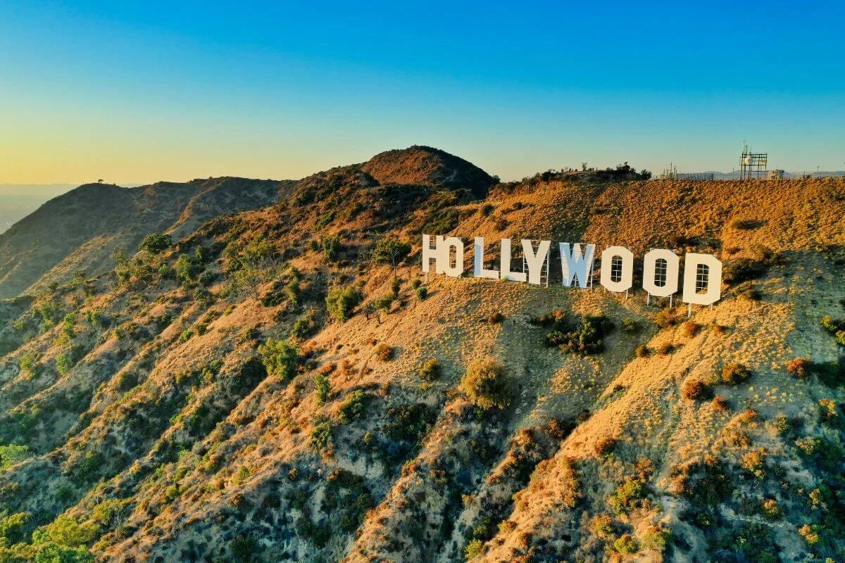 Aerial view of the iconic Hollywood sign on a sunlit hillside in Los Angeles with clear blue skies and surrounding mountains.