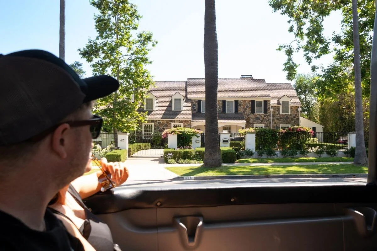 Man looking out from a tour vehicle window at a luxury home in Beverly Hills, surrounded by palm trees and manicured landscaping in Los Angeles.