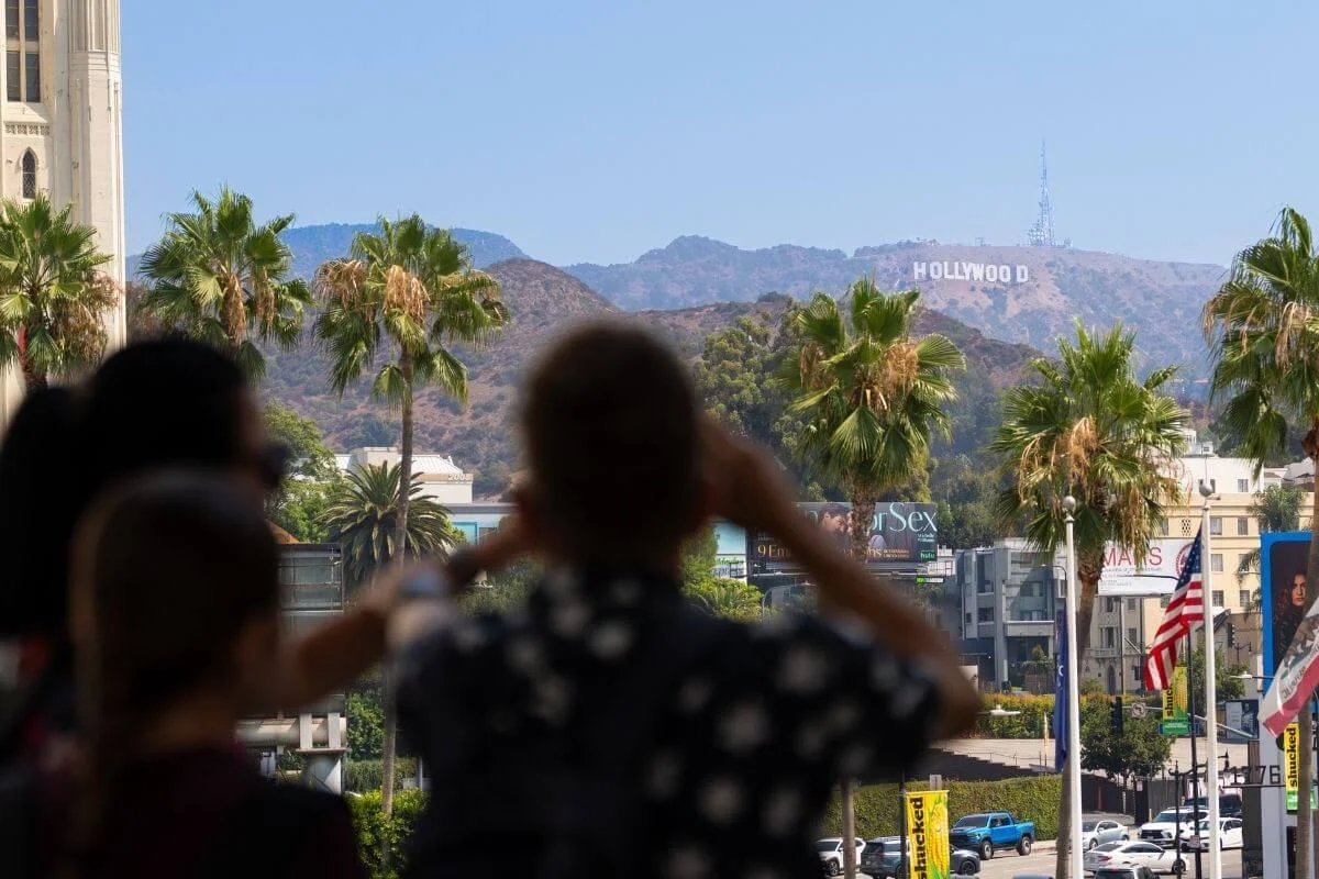 Tourists looking toward the Hollywood Sign in the hills above Los Angeles, with palm trees and city streets in the foreground.