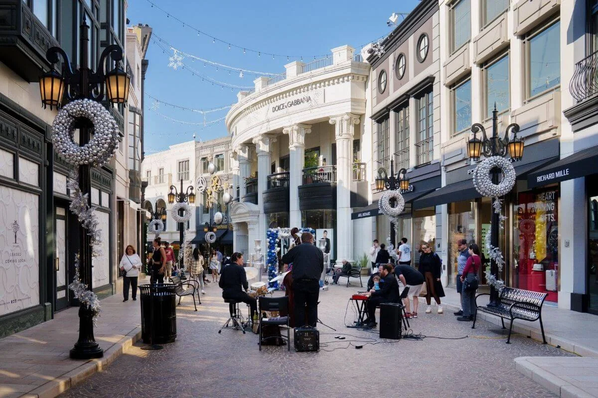 Street musicians performing live music along a decorated shopping street in Beverly Hills, with luxury stores and holiday wreaths lining Rodeo Drive.