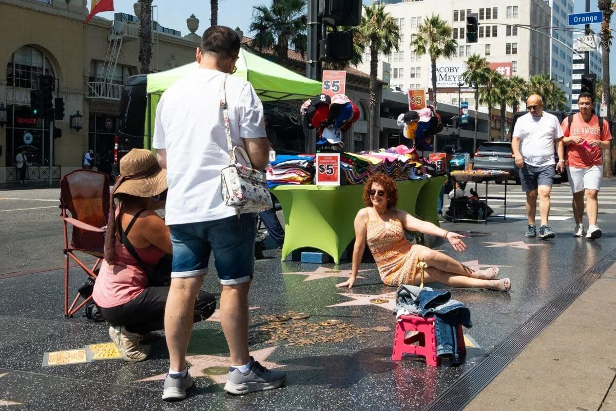 Visitors posing for photos on the Hollywood Walk of Fame, with street vendors and palm trees lining the busy Los Angeles sidewalk.