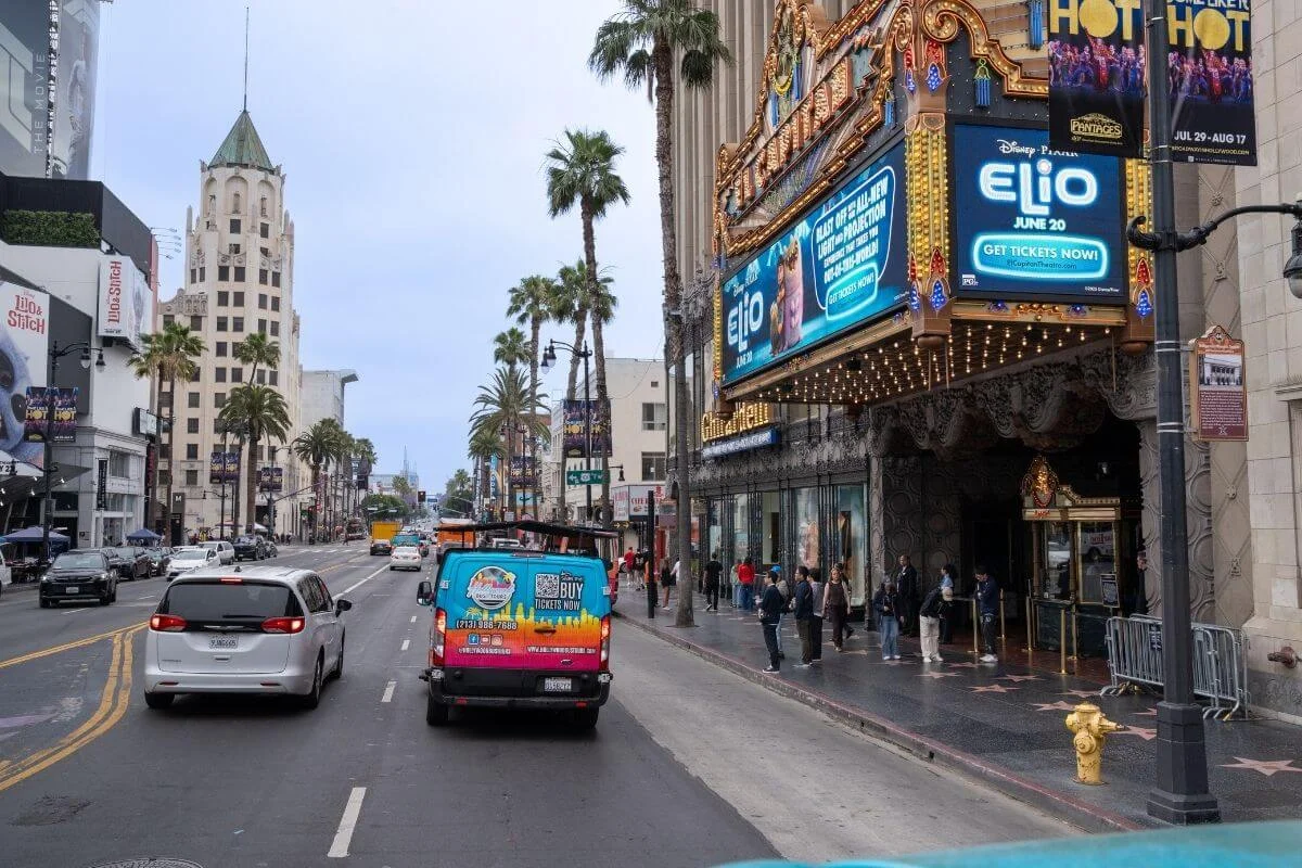 Street view of Hollywood Boulevard featuring El Capitan Theatre marquee, palm trees, traffic, and pedestrians along the Walk of Fame in Los Angeles.