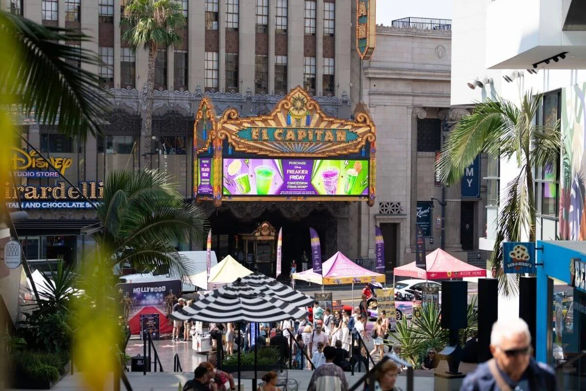 El Capitan Theatre marquee on Hollywood Boulevard surrounded by shops, palm trees, and tourists exploring the area in Los Angeles.