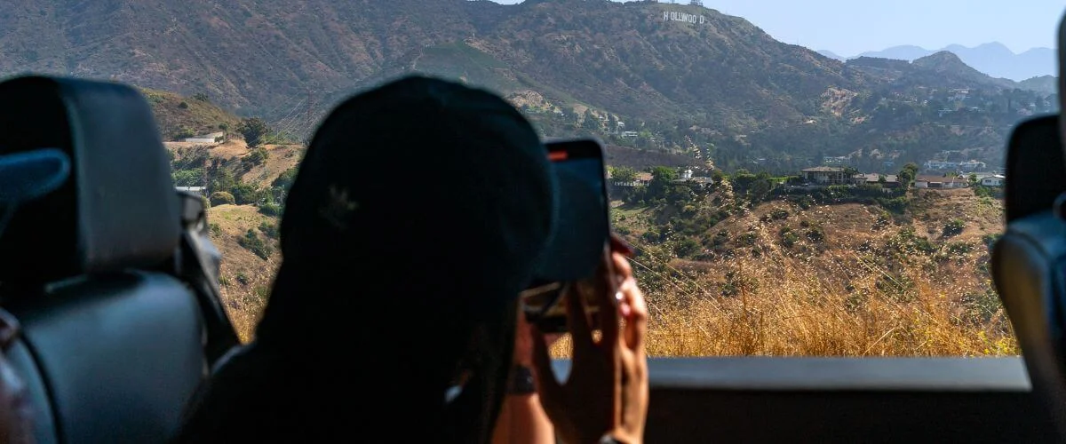 Person taking a photo of the Hollywood Hills from inside an open tour bus, with dry hillside terrain and scenic Los Angeles views in the background.