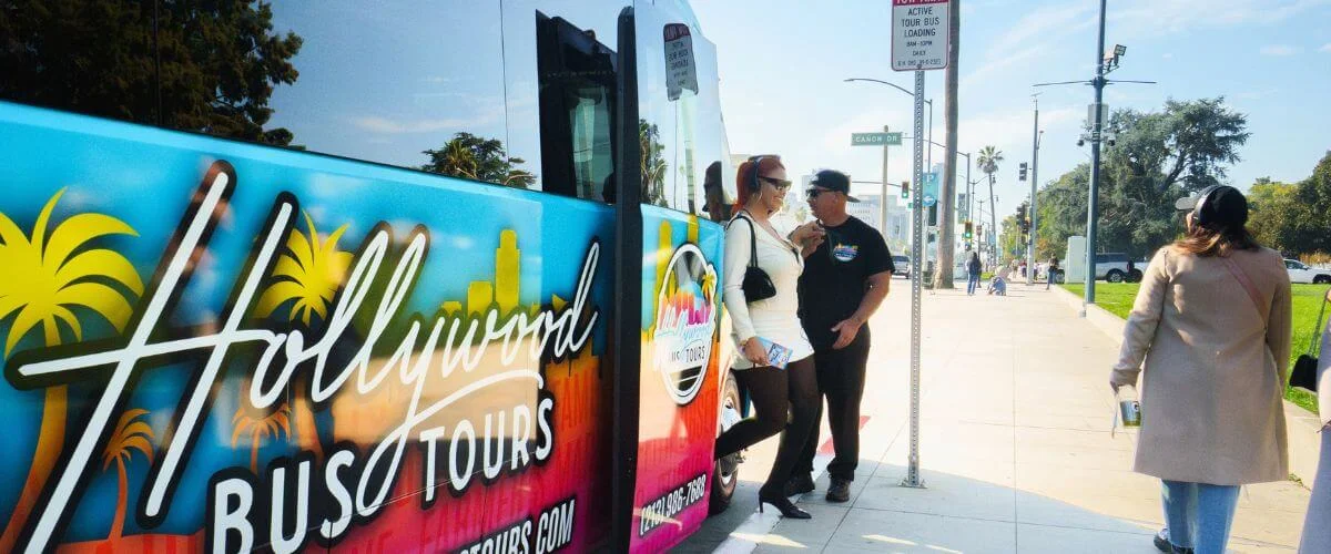 Tour guide assisting guests boarding a colorful Hollywood Bus Tours vehicle on a sunny Los Angeles street, showcasing a pickup experience.