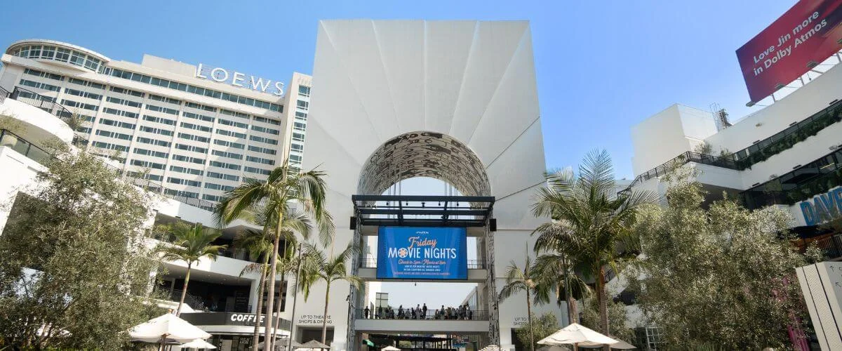 Main entrance courtyard at Ovation Hollywood with palm trees, shops, and Loews Hollywood Hotel in the background.
