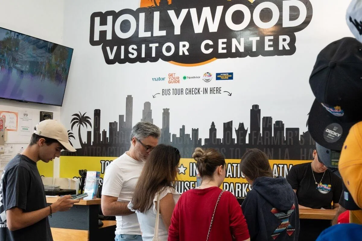 Visitors checking in at the Hollywood Visitor Center for bus tours, standing at the counter beneath signage showing tour partners and check-in information.