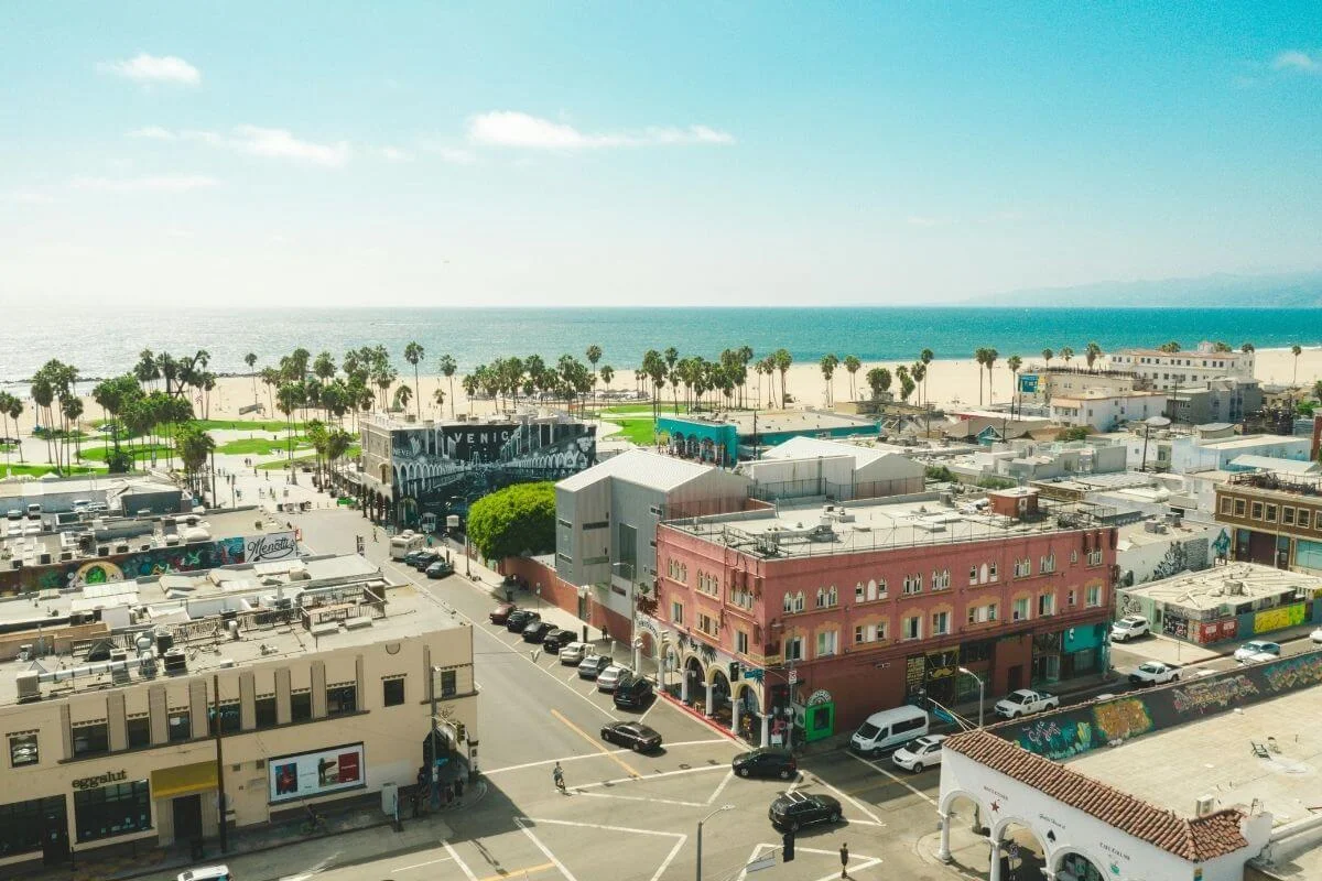 Aerial view of Venice Beach, Los Angeles, showing a grid of low-rise commercial buildings with colorful murals and the iconic Venice sign, leading to a palm-tree-lined boardwalk, sandy beach, and turquoise Pacific Ocean under a clear blue sky.