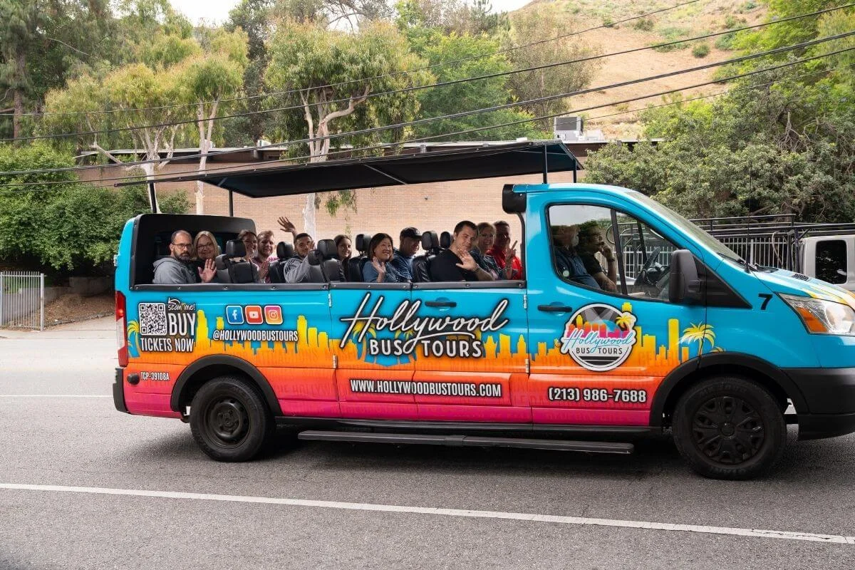 Tourists waving from an open-air Hollywood Bus Tours vehicle while sightseeing through Los Angeles on a sunny day.