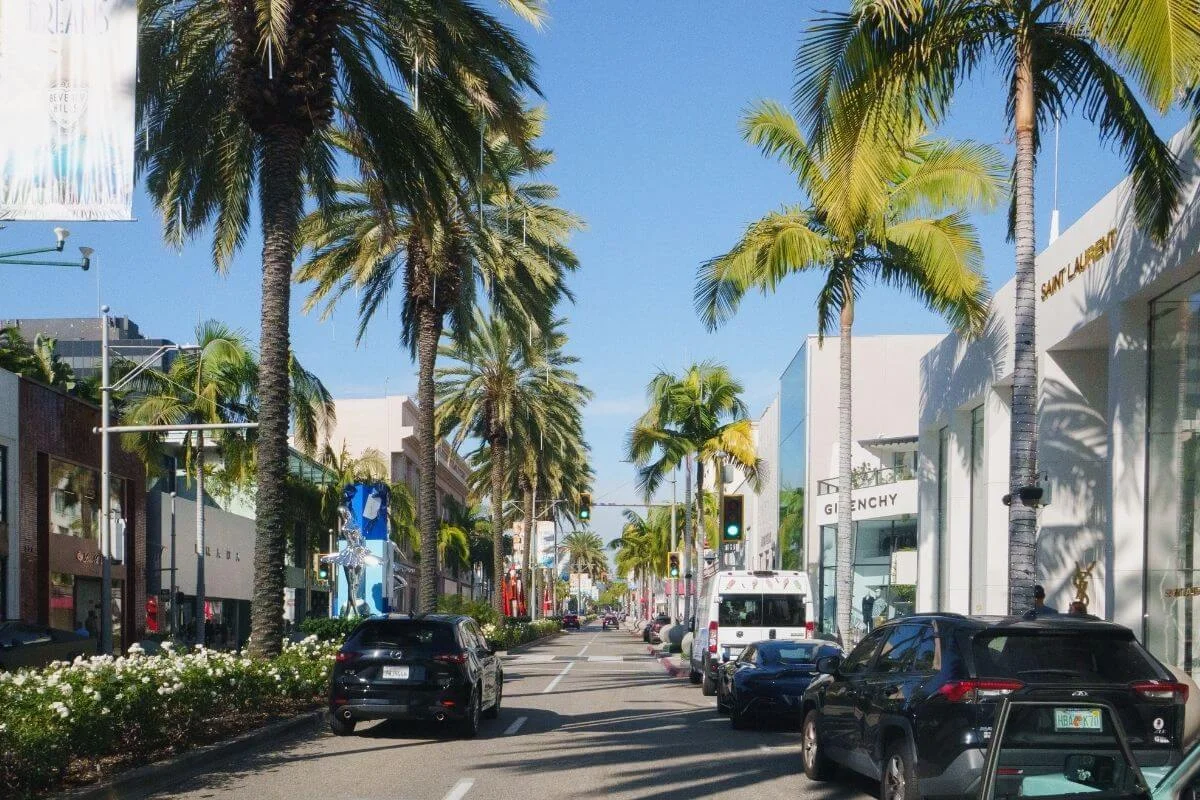 Street-level view of Rodeo Drive in Beverly Hills lined with tall palm trees and high-end boutiques including Givenchy and Saint Laurent on a clear sunny day.