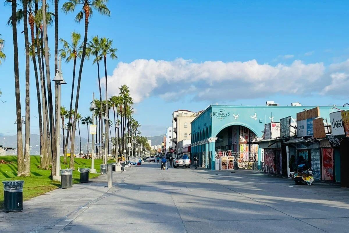 A wide, nearly empty Venice Beach boardwalk stretching into the distance, flanked by tall palm trees on the left and a row of closed vendor stalls and the teal-painted Big Daddy's Food Market on the right, under a bright blue sky with scattered clouds.