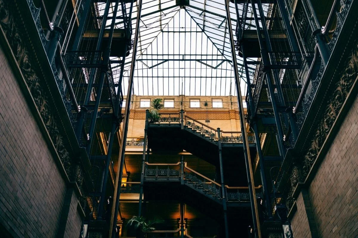 Looking up at the ornate Victorian interior of the Bradbury Building in downtown Los Angeles, showcasing its wrought iron staircases, open-air atrium, and glass skylight ceiling.