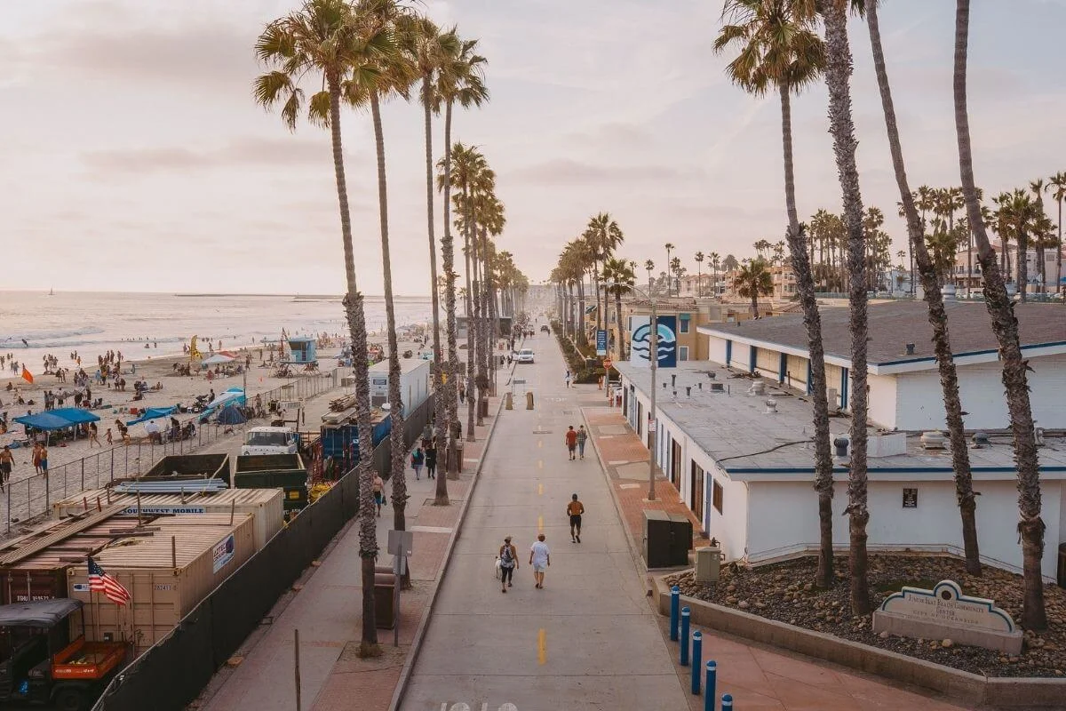 Elevated view of a wide beachside boardwalk lined with tall palm trees, with a busy sandy beach and ocean to the left and low-rise commercial buildings to the right, as pedestrians stroll under a warm hazy sunset sky
