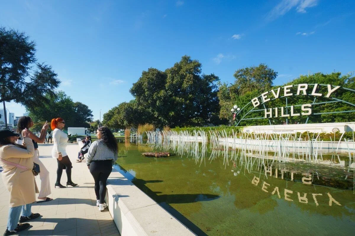 Tourists taking photos near the Beverly Hills sign and reflecting pool during a sunny afternoon in Los Angeles.
