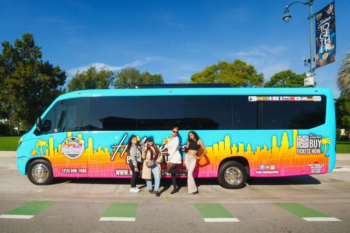 Four women posing in front of a vibrant teal Hollywood Bus Tours vehicle in Los Angeles, with the city skyline graphic and tour highlights printed along the side of the bus.