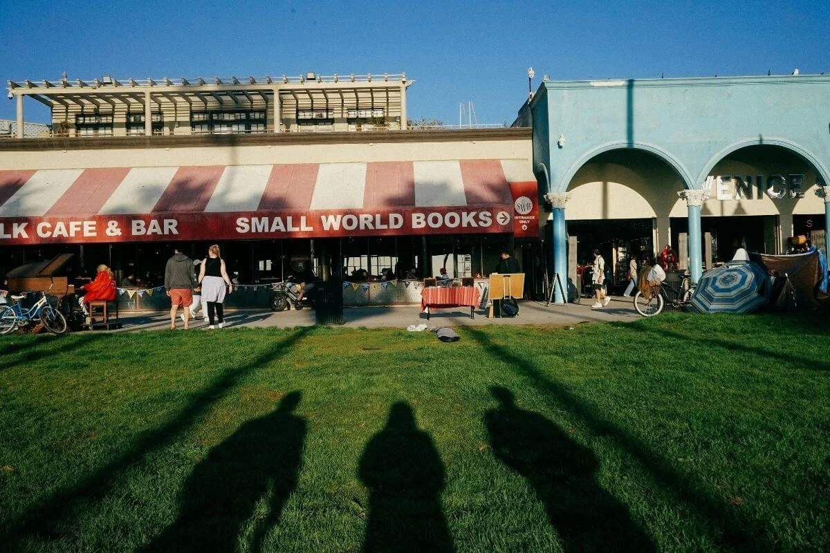 Storefront of Small World Books and an adjacent cafe and bar with a red and white striped awning on the Venice Beach boardwalk, with long shadows of onlookers cast across the green lawn in the foreground and the arched Venice sign visible to the right.