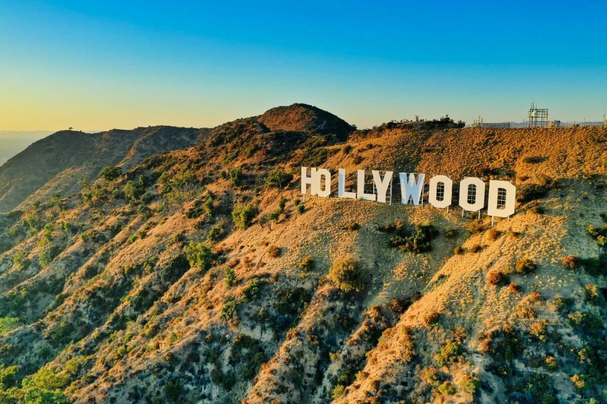 Aerial view of the iconic Hollywood Sign mounted on the dry golden hills of Los Angeles under a clear blue sky at golden hour.