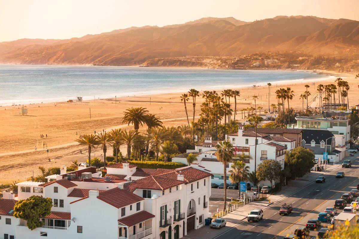 Scenic sunset view of Santa Monica Beach with palm trees, coastal homes, sandy shoreline, and mountains stretching along the California coast.