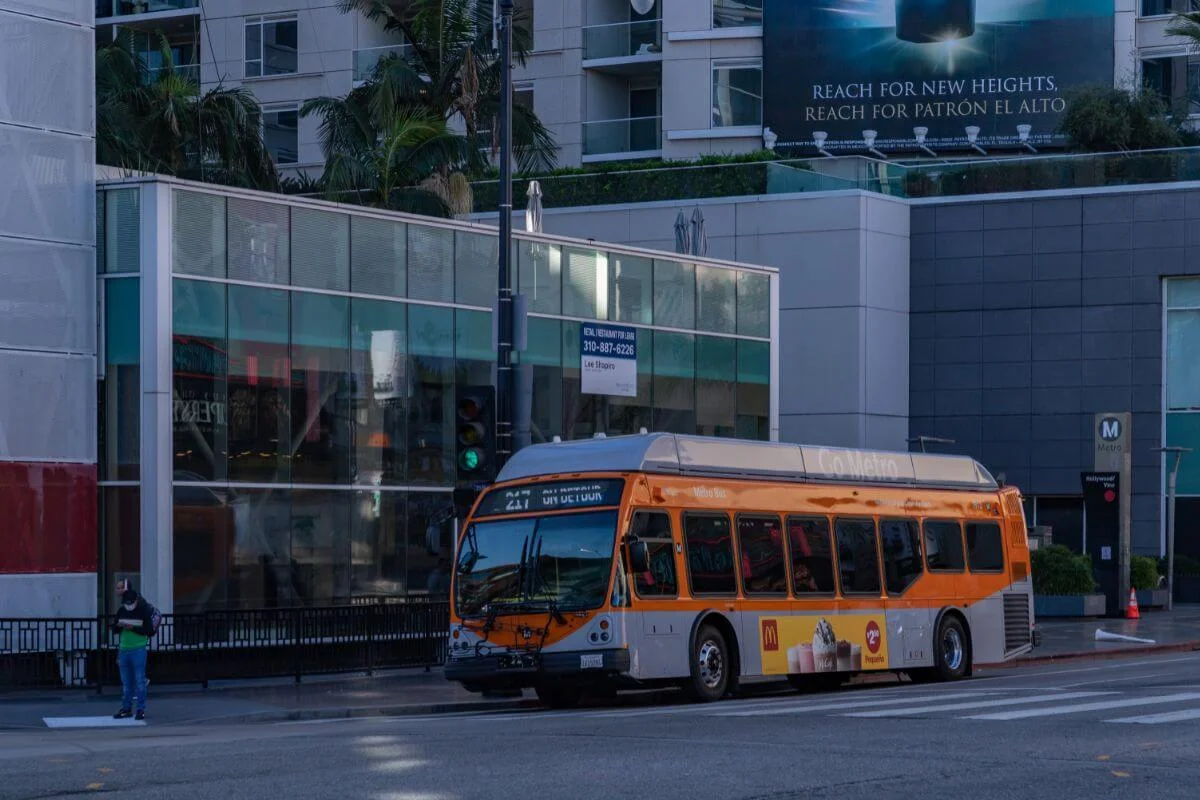 Orange Los Angeles Metro bus driving through downtown Los Angeles streets surrounded by modern buildings and city traffic.