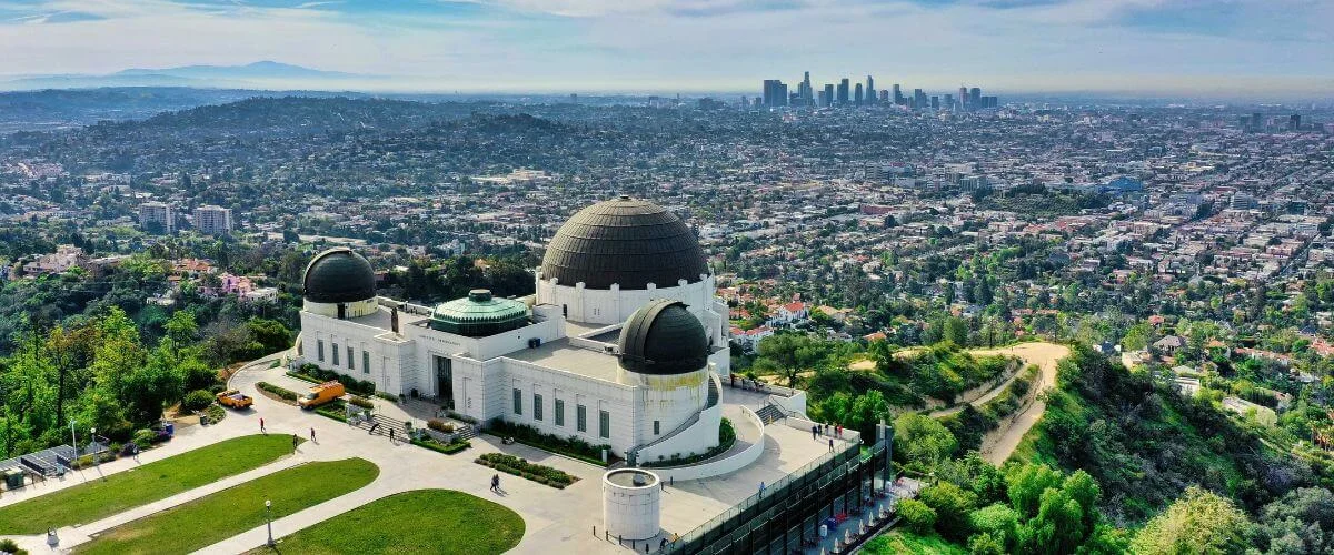Aerial view of Griffith Observatory in Los Angeles, with its iconic white domed building surrounded by green hills and the city skyline stretching into the background.